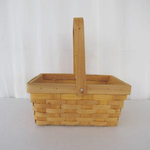 May include: A rectangular, light-colored woven basket with a tall, arched handle. The basket is made of thin, interwoven strips of wood, creating a textured surface. The handle is attached with a small wooden peg. Ideal for storage or display.