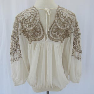 Puede incluir: Blusa color crema con mangas largas y bordados marrones intrincados en los hombros y el pecho. La blusa tiene un escote en V y una cintura fruncida, con mangas recogidas en las mu&ntilde;ecas.