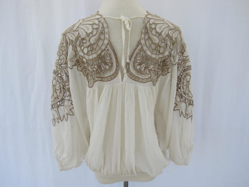 Puede incluir: Blusa color crema con mangas largas y bordados marrones intrincados en los hombros y el pecho. La blusa tiene un escote en V y una cintura fruncida, con mangas recogidas en las mu&ntilde;ecas.