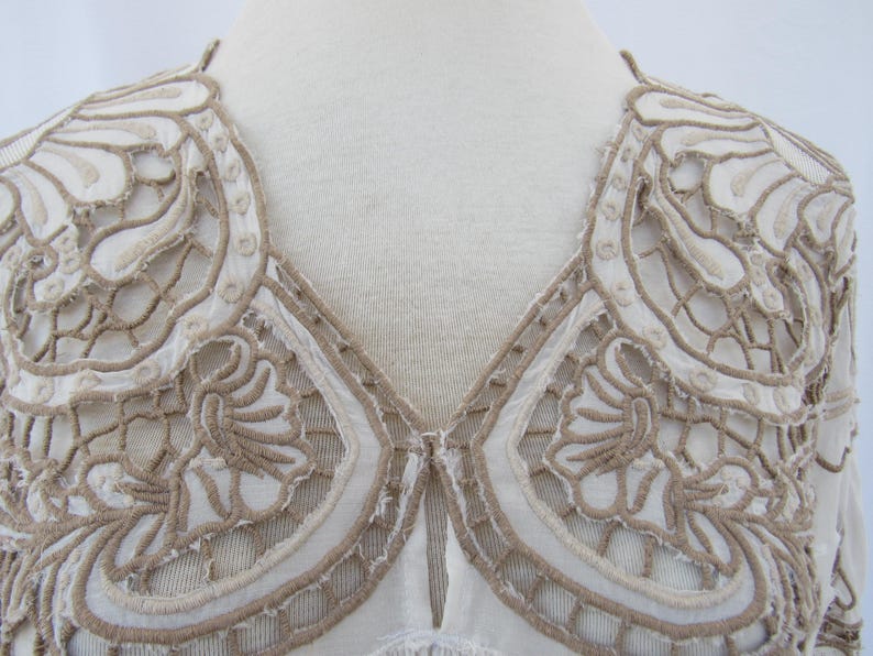 Puede incluir: Primer plano de una blusa color crema con intrincados bordados calados en un tono marr&oacute;n contrastante. El bordado forma un patr&oacute;n decorativo alrededor del escote y los hombros, creando un dise&ntilde;o delicado y texturizado. La blusa se exhibe en un maniqu&iacute;.