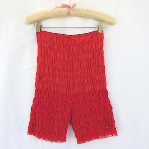 May include: Bright red shorts with a textured, ruffled design. The shorts are hanging on a padded hanger with a pink bow. The fabric has a crinkled appearance, and the shorts have a lace trim at the bottom.