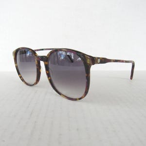 May include: Vintage sunglasses with a tortoiseshell pattern frame. The lenses are a gradient gray, transitioning from light at the bottom to dark at the top. The arms are thin and curve down to the ear. The sunglasses are set against a white background.