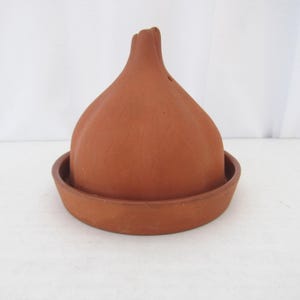 May include: A terracotta cloche with a rounded top and a circular base. The cloche is a warm, earthy brown color and appears to be made of unglazed clay. It is designed to sit on the base, creating a dome shape.