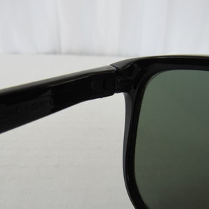 May include: Close-up of the hinge of a pair of black sunglasses with green lenses. The sunglasses are made of plastic and have a simple design.