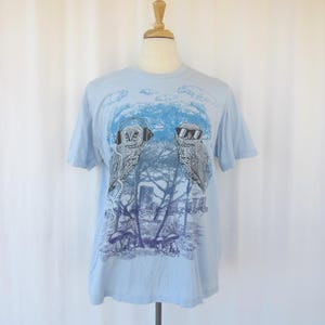 May include: Light blue t-shirt featuring a graphic of two owls. One owl wears headphones, the other sunglasses. The design includes trees, mushrooms, and a blue sky background. The shirt is displayed on a mannequin.
