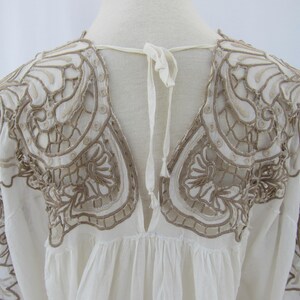 Puede incluir: Una blusa blanca con intrincados detalles bordados en color marr&oacute;n claro. El bordado se concentra alrededor de los hombros y el escote, con un cierre de lazo en la espalda. Las mangas son largas y la tela parece ligera.