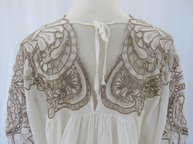Puede incluir: Una blusa blanca con intrincados detalles bordados en color marr&oacute;n claro. El bordado se concentra alrededor de los hombros y el escote, con un cierre de lazo en la espalda. Las mangas son largas y la tela parece ligera.