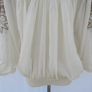 Puede incluir: Blusa color crema con mangas largas y bordados marrones intrincados en los hombros. La blusa tiene cintura y mangas fruncidas, creando una silueta fluida. La tela parece ligera y ligeramente transparente.