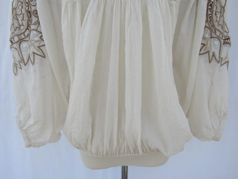 Puede incluir: Blusa color crema con mangas largas y bordados marrones intrincados en los hombros. La blusa tiene cintura y mangas fruncidas, creando una silueta fluida. La tela parece ligera y ligeramente transparente.