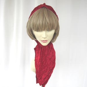 May include: A red velvet headband and scarf set. The headband is twisted at the top, and the scarf hangs down. The set is displayed on a mannequin head with blonde hair and a fair complexion. The background is white.