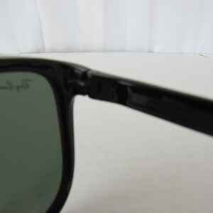 May include: Close-up of the black frame of a pair of sunglasses with green lenses. The sunglasses have the text "Ray-Ban" printed on the frame.