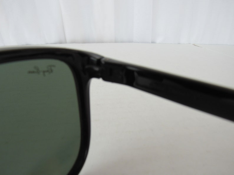 May include: Close-up of the black frame of a pair of sunglasses with green lenses. The sunglasses have the text "Ray-Ban" printed on the frame.