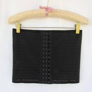 May include: A black waist trainer belt featuring a perforated mesh fabric design. The garment includes a central vertical panel with multiple rows of metal hook-and-eye closures for adjustable fastening, suspended from a padded beige hanger.