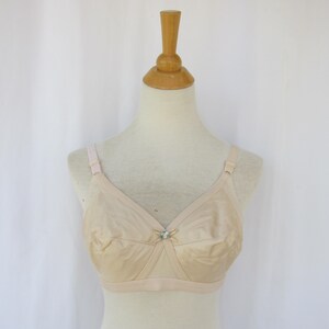 May include: A beige brassiere featuring a smooth, satin-like fabric finish, adjustable shoulder straps, and a small decorative center bow detail. The garment is displayed on a mannequin, showcasing a full-coverage design with structured cups.