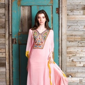 May include: A long, pink, flowing dress with colourful embroidery and yellow fringe. The dress is being worn by a person standing in front of a wooden door.