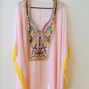 May include: A pink kaftan with yellow fringe trim and colourful embroidery on the neckline. The embroidery features a floral design with birds and geometric patterns.