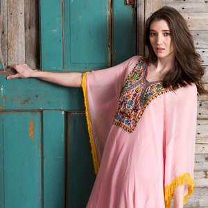 May include: A woman wearing a pink, long-sleeved kaftan with colourful embroidery and yellow fringe. She is standing in front of a weathered wooden door.