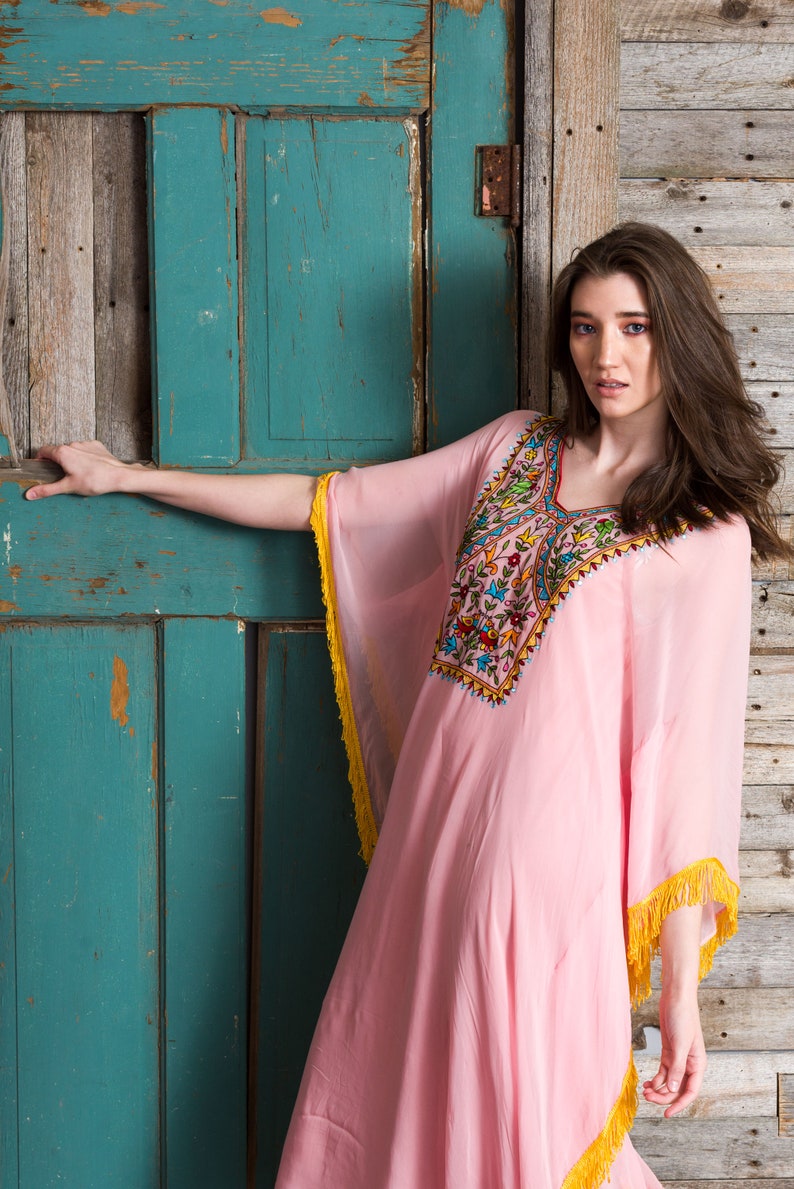 May include: A woman wearing a pink, long-sleeved kaftan with colourful embroidery and yellow fringe. She is standing in front of a weathered wooden door.
