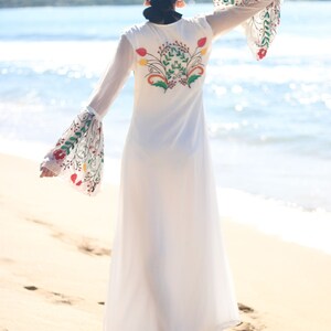 Peut inclure: Une robe longue blanche avec des broderies florales au dos et sur les manches. La robe a de longues manches transparentes avec des poignets &eacute;vas&eacute;s. Le mod&egrave;le est sur une plage de sable avec la mer en arri&egrave;re-plan.