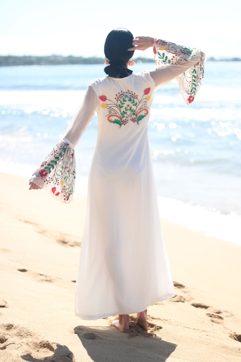 Peut inclure: Une robe longue blanche avec des broderies florales au dos et sur les manches. La robe a de longues manches transparentes avec des poignets &eacute;vas&eacute;s. Le mod&egrave;le est sur une plage de sable avec la mer en arri&egrave;re-plan.