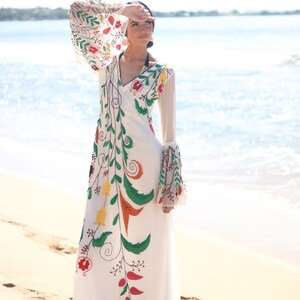 Peut inclure: Une longue robe blanche avec des broderies florales color&eacute;es. La robe pr&eacute;sente un col en V, de longues manches transparentes et une jupe fluide. La broderie comprend des motifs floraux rouges, verts, jaunes et oranges. La robe est port&eacute;e sur une plage de sable avec l'oc&eacute;an en arri&egrave;re-plan.