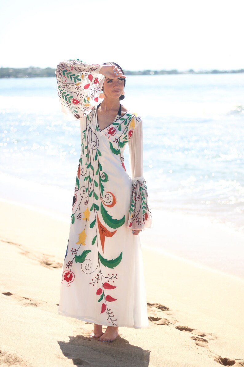 Peut inclure: Une longue robe blanche avec des broderies florales color&eacute;es. La robe pr&eacute;sente un col en V, de longues manches transparentes et une jupe fluide. La broderie comprend des motifs floraux rouges, verts, jaunes et oranges. La robe est port&eacute;e sur une plage de sable avec l'oc&eacute;an en arri&egrave;re-plan.