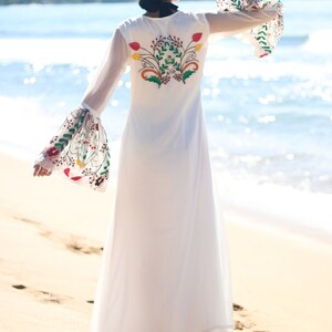 Peut inclure: Longue robe blanche avec broderie florale au dos et sur les manches. La robe a de longues manches cloches transparentes avec des motifs floraux color&eacute;s. La robe est port&eacute;e sur une plage de sable avec l'oc&eacute;an en arri&egrave;re-plan.