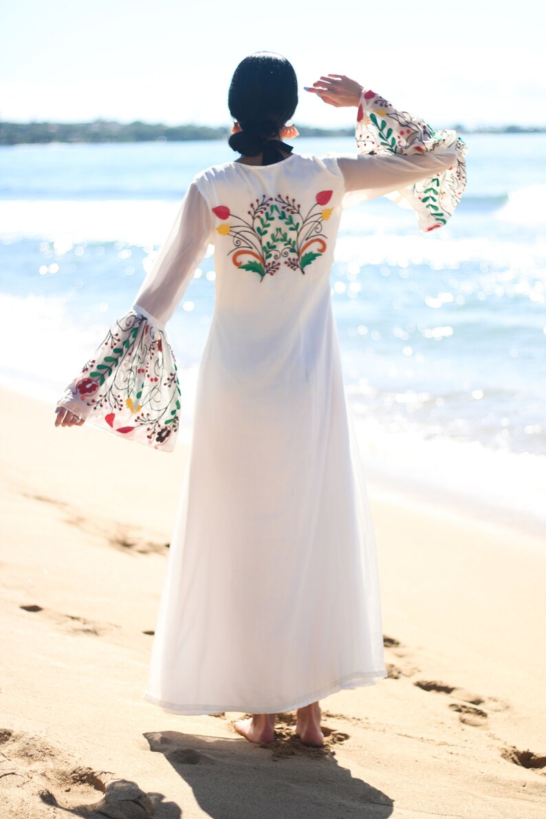 Peut inclure: Longue robe blanche avec broderie florale au dos et sur les manches. La robe a de longues manches cloches transparentes avec des motifs floraux color&eacute;s. La robe est port&eacute;e sur une plage de sable avec l'oc&eacute;an en arri&egrave;re-plan.