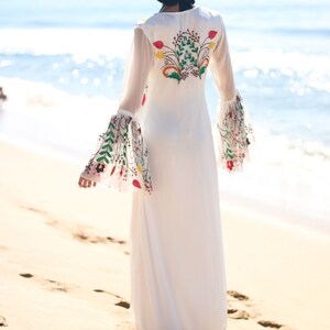 Peut inclure: Une robe longue blanche avec de longues manches transparentes et des broderies florales au dos et sur les manches. La robe est port&eacute;e sur une plage de sable avec l'oc&eacute;an en arri&egrave;re-plan. La broderie pr&eacute;sente des fleurs rouges, vertes et jaunes.