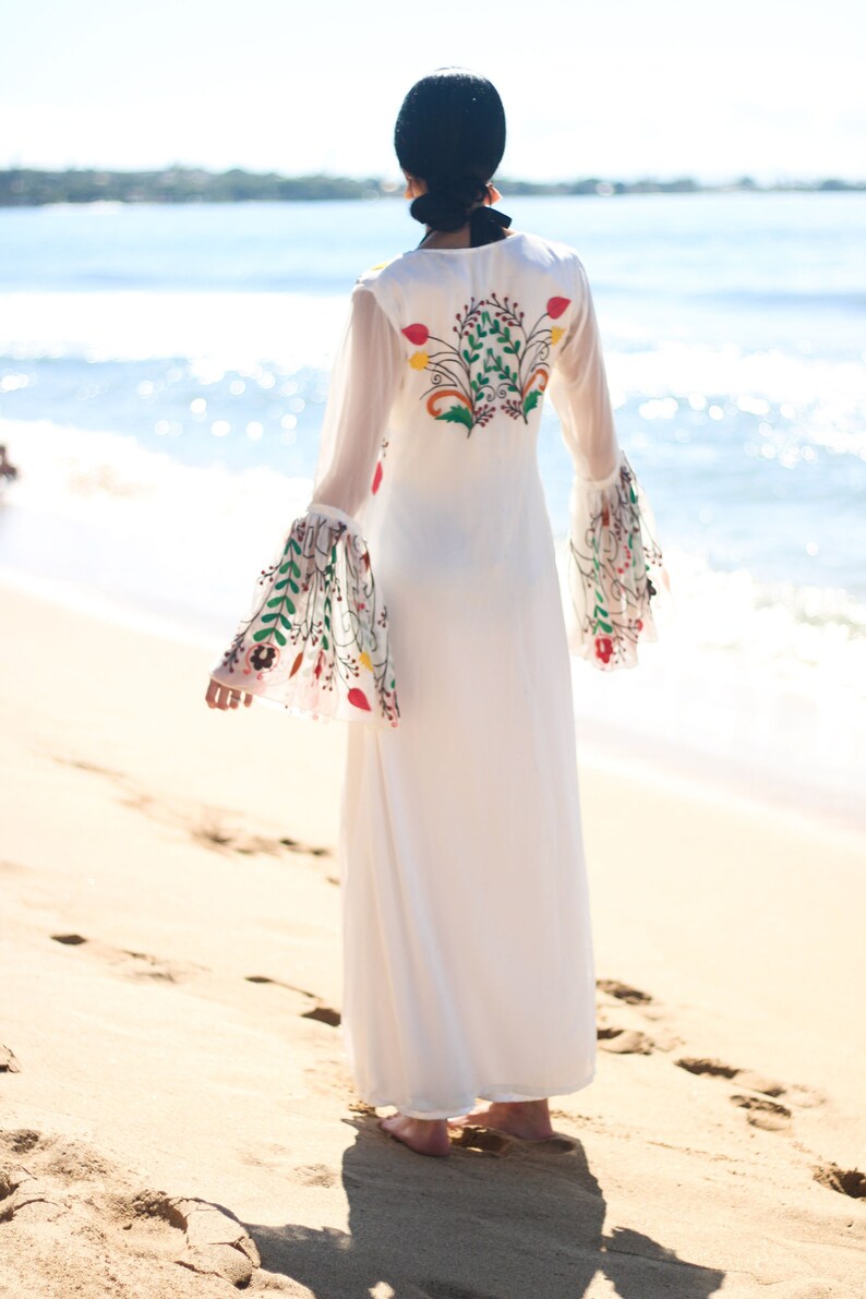 Peut inclure: Une robe longue blanche avec de longues manches transparentes et des broderies florales au dos et sur les manches. La robe est port&eacute;e sur une plage de sable avec l'oc&eacute;an en arri&egrave;re-plan. La broderie pr&eacute;sente des fleurs rouges, vertes et jaunes.