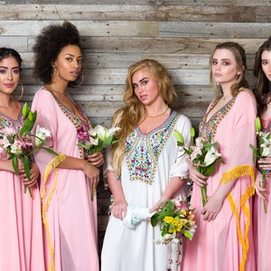May include: Five women wearing long, pink and white dresses with colourful embroidery and gold fringe. They are holding bouquets of flowers and standing in front of a wood panel wall.