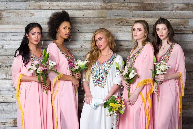 May include: Five women wearing long, pink and white dresses with colourful embroidery and gold fringe. They are holding bouquets of flowers and standing in front of a wood panel wall.