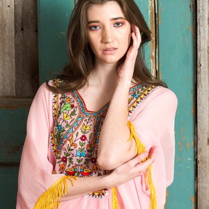May include: A pink embroidered kaftan with yellow fringe trim. The kaftan features a floral and geometric design in shades of blue, green, red, and yellow.