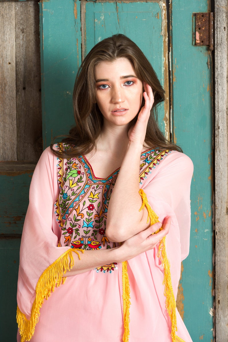 May include: A pink embroidered kaftan with yellow fringe trim. The kaftan features a floral and geometric design in shades of blue, green, red, and yellow.