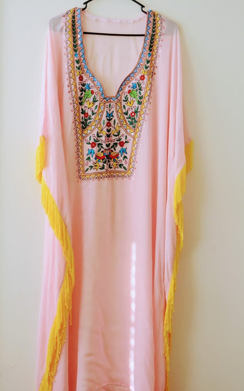 May include: A pink kaftan with yellow fringe trim and colourful embroidery on the neckline. The embroidery features a floral design with birds and geometric patterns.