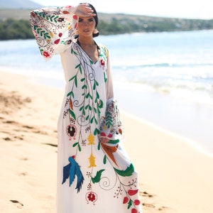 Peut inclure: Une robe blanche &agrave; manches longues avec un col en V et des broderies florales et d'oiseaux color&eacute;es. La robe pr&eacute;sente des manches &eacute;vas&eacute;es et est port&eacute;e sur une plage de sable avec l'oc&eacute;an en arri&egrave;re-plan. La broderie comprend des accents rouges, verts, bleus et jaunes.