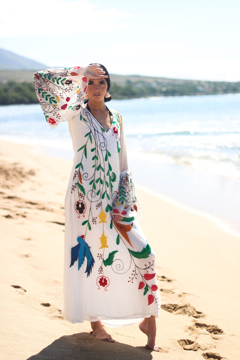 Peut inclure: Une robe blanche &agrave; manches longues avec un col en V et des broderies florales et d'oiseaux color&eacute;es. La robe pr&eacute;sente des manches &eacute;vas&eacute;es et est port&eacute;e sur une plage de sable avec l'oc&eacute;an en arri&egrave;re-plan. La broderie comprend des accents rouges, verts, bleus et jaunes.