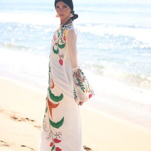 Peut inclure: Une robe longue blanche avec des broderies florales color&eacute;es. La robe pr&eacute;sente de longues manches transparentes et une silhouette fluide. La broderie comprend des feuilles vertes, des baies rouges et des accents orange. La robe est port&eacute;e sur une plage de sable.