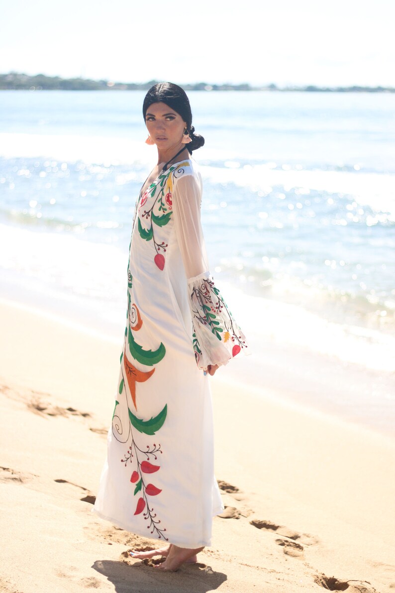 Peut inclure: Une robe longue blanche avec des broderies florales color&eacute;es. La robe pr&eacute;sente de longues manches transparentes et une silhouette fluide. La broderie comprend des feuilles vertes, des baies rouges et des accents orange. La robe est port&eacute;e sur une plage de sable.