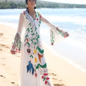 Peut inclure: Longue robe blanche orn&eacute;e de broderies florales et d'oiseaux color&eacute;s. La robe pr&eacute;sente un col en V, de longues manches transparentes et une jupe fluide. Les broderies comprennent des accents verts, rouges, jaunes et bleus, cr&eacute;ant un style boh&egrave;me.