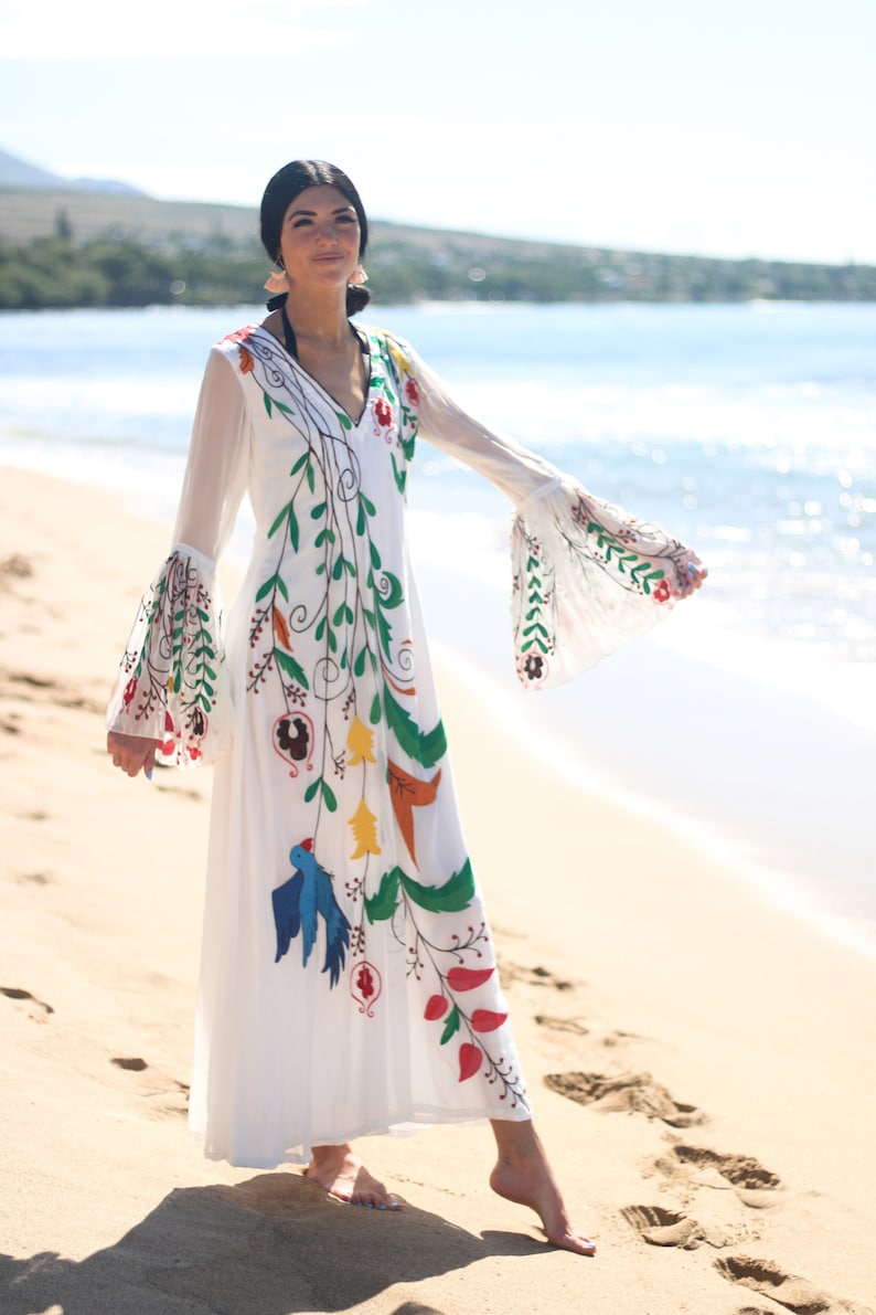 Peut inclure: Longue robe blanche orn&eacute;e de broderies florales et d'oiseaux color&eacute;s. La robe pr&eacute;sente un col en V, de longues manches transparentes et une jupe fluide. Les broderies comprennent des accents verts, rouges, jaunes et bleus, cr&eacute;ant un style boh&egrave;me.