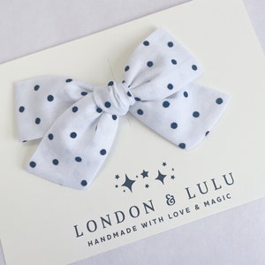 May include: White fabric bow with black polka dots. The bow is tied with a knot in the center. The bow is on a white background with the text "LONDON & LULU HANDMADE WITH LOVE & MAGIC" in black.