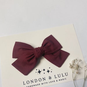 May include: A burgundy fabric bow with a simple design. The bow is handmade and has a tag that reads "London & Lulu Handmade with Love & Magic".