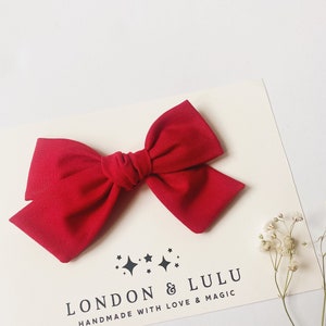 May include: A large red fabric bow with a center knot. The bow is on a white background with a small floral sprig and a logo that reads "LONDON & LULU HANDMADE WITH LOVE & MAGIC".