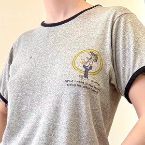 May include: Gray ringer t-shirt with black trim and a graphic. The graphic is a cartoon of a person holding a drink and the text "What I need is for people telling me what I need".