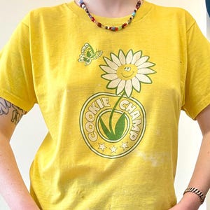 May include: Yellow t-shirt with a daisy graphic and the text "COOKIE CHAMP". The shirt also features a butterfly graphic. The shirt is paired with blue jeans.