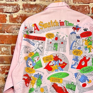 May include: Pink button-up shirt with a comic strip design on the back. The comic strip features colorful illustrations of characters, castles, and the text "A Swatch in Time". The shirt is hanging on a wooden hanger.