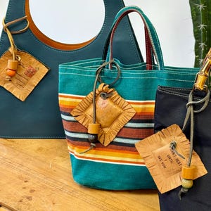 May include: Three stylish handbags in teal, turquoise, and black sit on a wooden surface. The teal bag has a large circular handle, while the turquoise bag features colorful stripes. The black bag has a bamboo handle and the text "ESPRIT DE CORP". Each bag has a decorative tag.