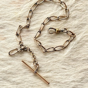 May include: A vintage gold-toned pocket watch chain with a T-bar and a clasp. The chain is composed of oval links and is displayed on a textured, off-white surface.