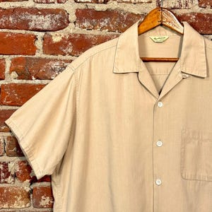 May include: A tan short-sleeved button-down shirt with a camp collar, hanging on a wooden hanger. The shirt has a single chest pocket and white buttons. The background is a weathered brick wall.